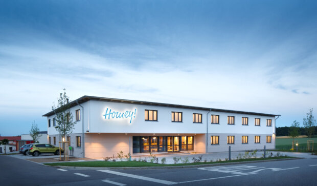 Außenansicht des modernen Homey! MicroApartment-Gebäudes in der Abenddämmerung mit beleuchtetem Eingangsbereich und stilvoller Architektur.